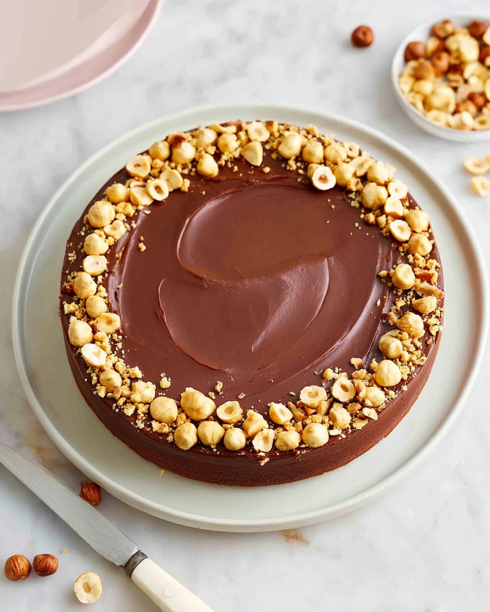 A round cake with three visible layers sits on a white plate on a white marbled surface. The bottom layer is a thin, golden-brown crust. The middle layer is thick and light brown with a smooth, creamy texture. The top layer is a glossy, dark chocolate ganache spread evenly across the surface with some gentle swirls visible. Around the edge of the top layer, there is a border of halved and chopped toasted hazelnuts, golden and slightly browned. A silver fork and knife with white handles rest nearby, adding a simple, elegant touch. Photo taken with an iphone --ar 4:5 --v 7