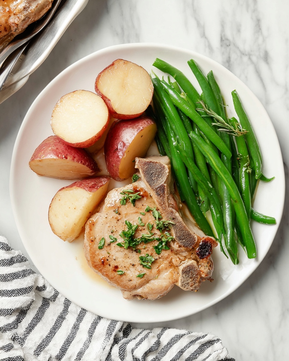 A white plate holds three main parts: a large piece of lightly browned pork chop with some green herb bits on top in the lower right area, smooth green beans with a fresh glossy look arranged in a neat bundle along the right edge above the chop, and red-skinned potatoes quartered and stacked in the upper left quadrant showing creamy off-white inside. The plate sits on a white marbled surface with a striped cloth nearby. photo taken with an iphone --ar 4:5 --v 7