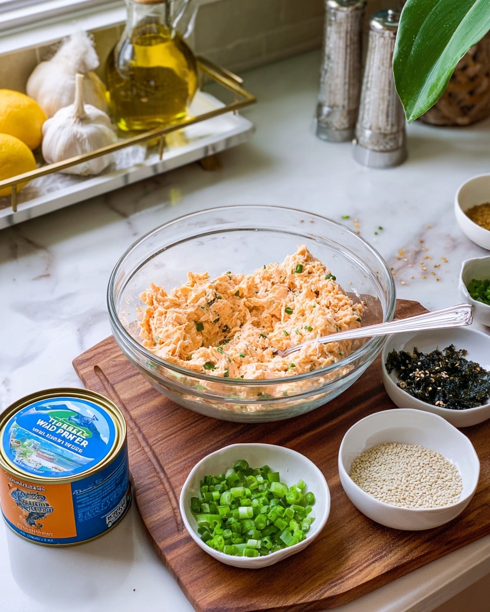 A clear glass bowl filled with a light orange chunky mixture, likely tuna salad, sits in the center of a wooden board. A metal spoon is partially placed inside the bowl resting on the right side. Surrounding the bowl are three small white bowls: one filled with chopped green onions, another with white sesame seeds, and the last one containing seaweed pieces mixed with sesame seeds and spices. A blue and orange can of Wild Planet tuna is placed in front of the glass bowl. The background features a white marbled counter with a white tray holding a clear glass bottle of olive oil, a garlic bulb, and lemons. There is also a silver container, a green plant leaf, and white salt and pepper shakers on the white marbled countertop. Photo taken with an iphone --ar 4:5 --v 7